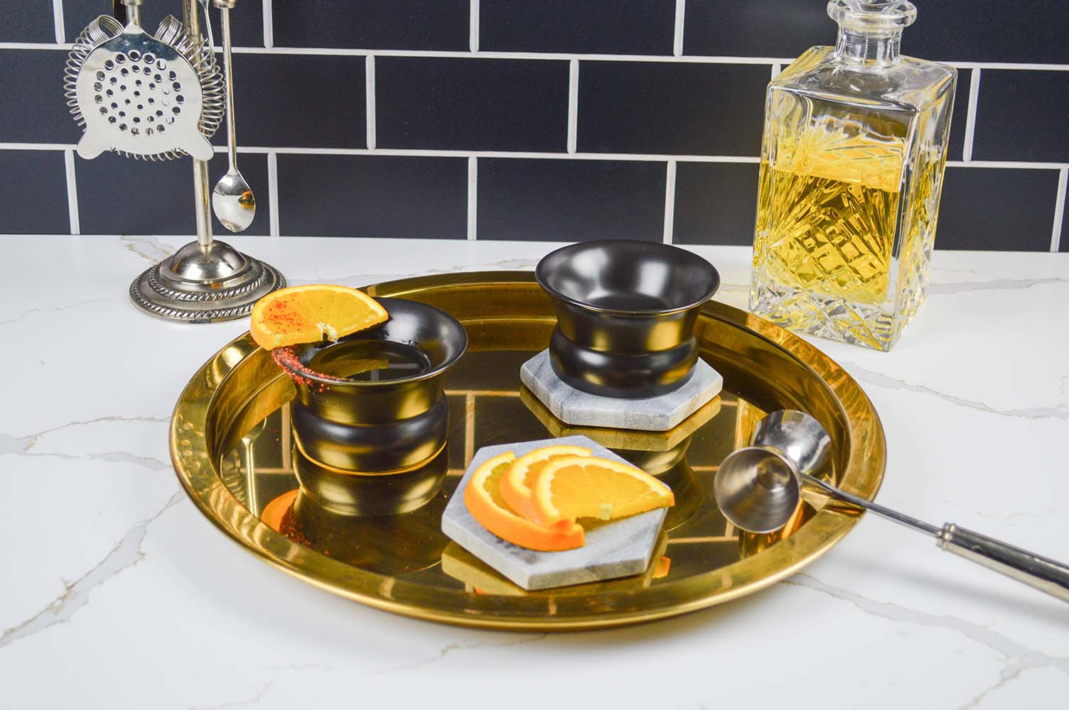 Two volcanic black Oaxaca glasses on a bronze circular tray, surrounded by bar ware and orange slices.