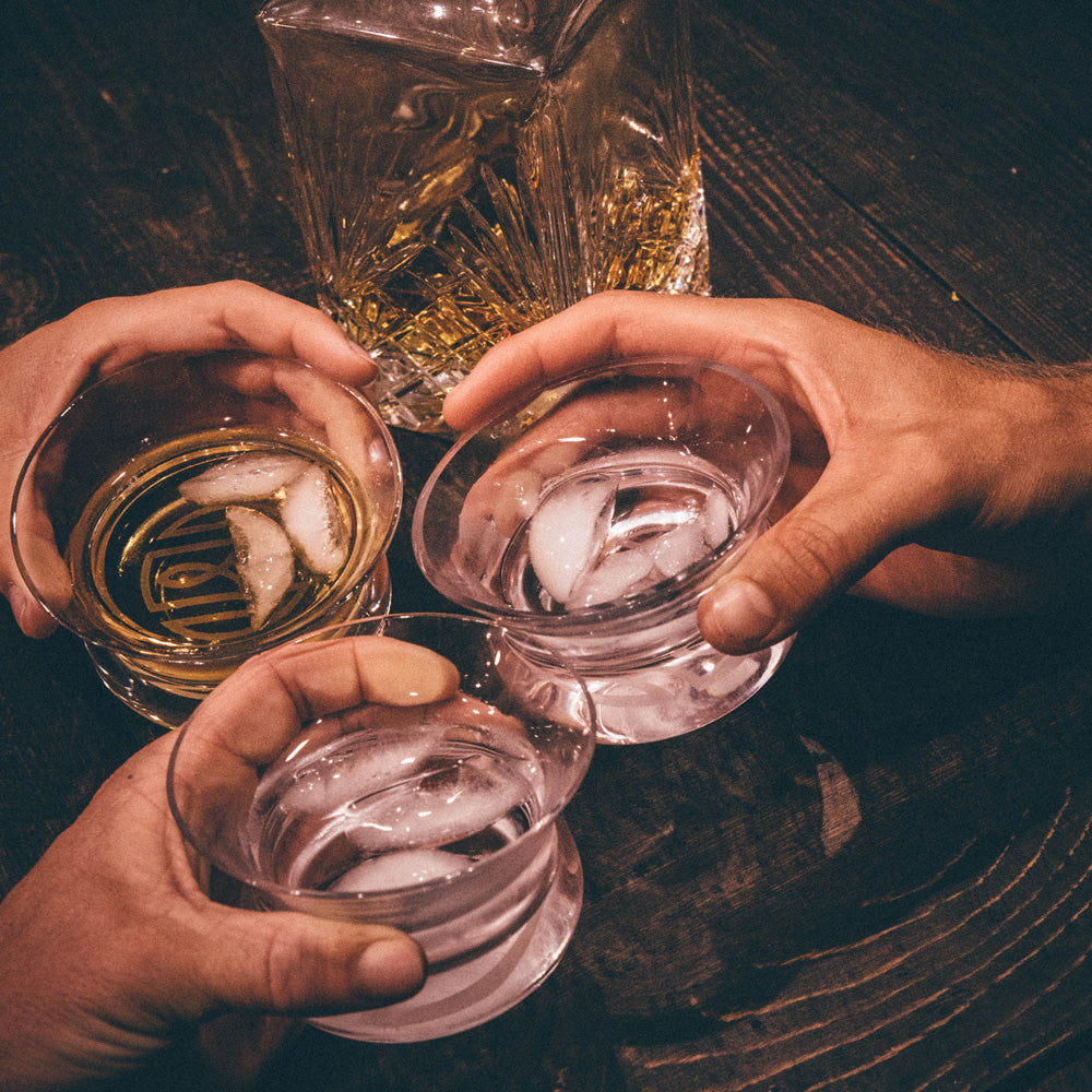 Three Oaxaca glasses being cheersed over a wooden table, each filled with tequila and ice.