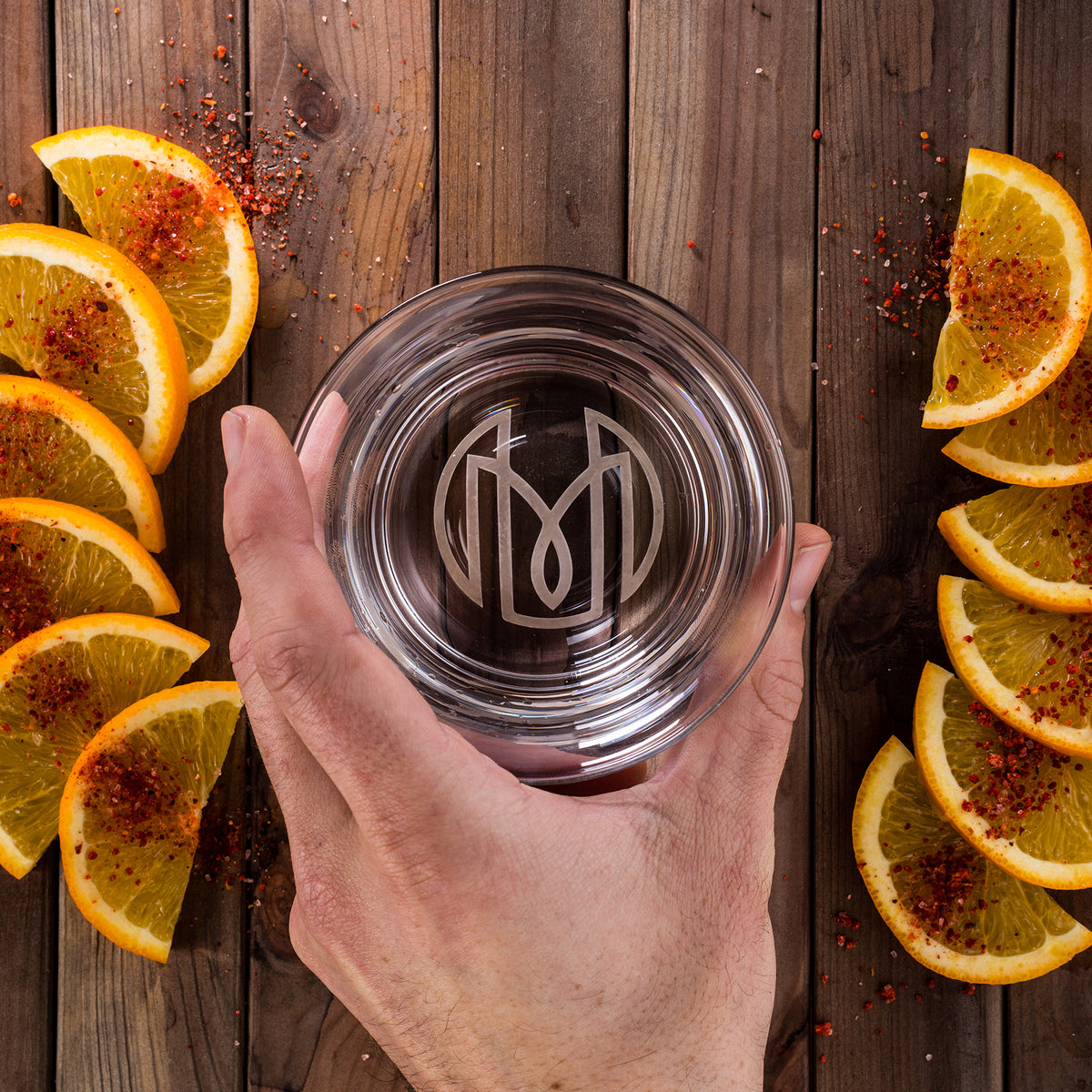 An overhead shot of the Oaxaca glass showing the Historically Modern monogram. It's on being placed down onto a wood table, surrounded by arcs of orange slices on either side, with sprinked red salt.
