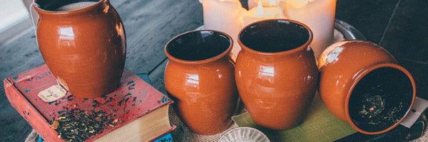 Four Jalisco jugs sitting on a wooden table with lit candles in the background. One of the jugs sits on stacked red and blue books with loose leaf tea sprinked around. The fourth jug is laying on its side with loose leaf tea inside.