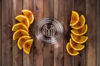 Overhead shot of the Oaxaca glass showing the Historically Modern monogram on the bottom of the glass. The glass is on a wood table surrounded by two arcs of orange slices covered in red salt.