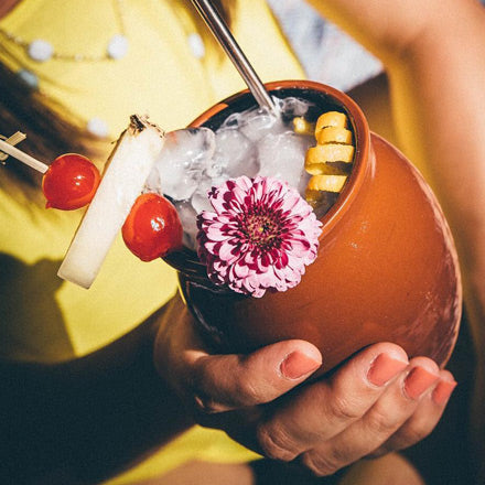 A Jalisco jug filled with a piña colada held by a lady in yellow clothing. The Jalisco jug is garnished with skewered cherries, pineapple, a citrus twist, and a flower.