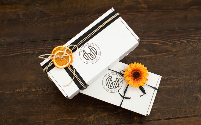 Two white cardboard Oaxaca glass boxes stacked on each other on a wood table. The boxes are wrapped with ribbon and twine with decorations of dried oranges and sunflowers.