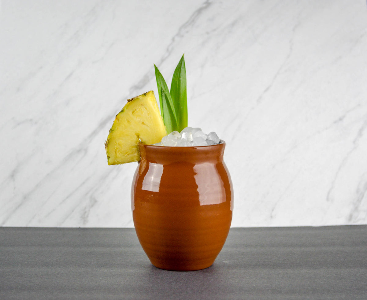 A Glossy finish Jalisco jug on a concrete countertop with a marble background. The jug is filled with small ice cubes and garnished with a pineapple slice and pineapple leaves.