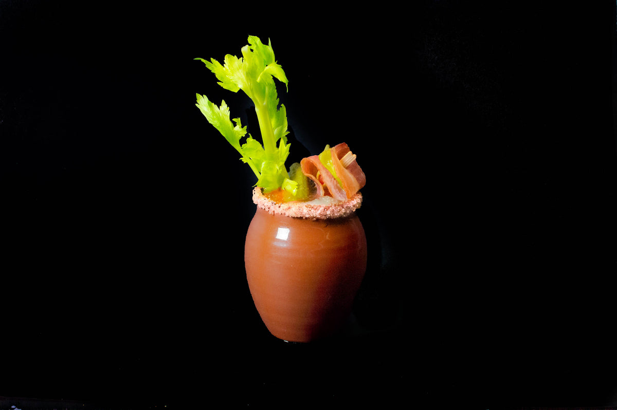 A glossy finish Jalisco jug on a black background. It's heavily rimmed with salt, filled with a Bloody Mary cocktail, and garnished with a celery stalk and skewered bacon, olives, and pickles.