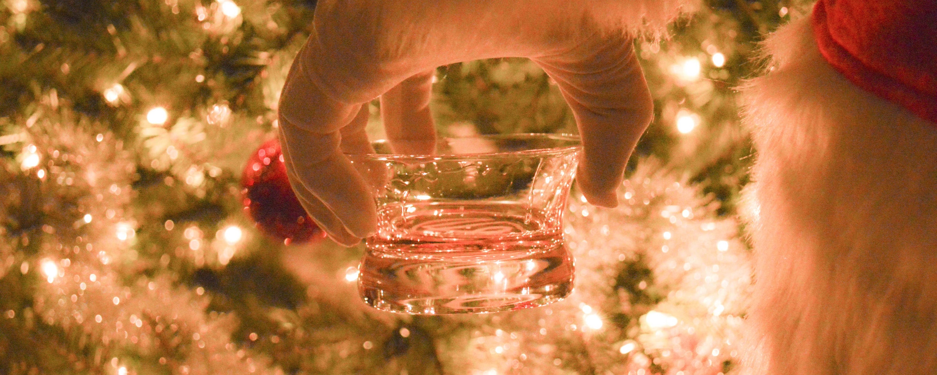 Close up of Santa's hand holding a Oaxaca glass in front of a lit Christmas tree