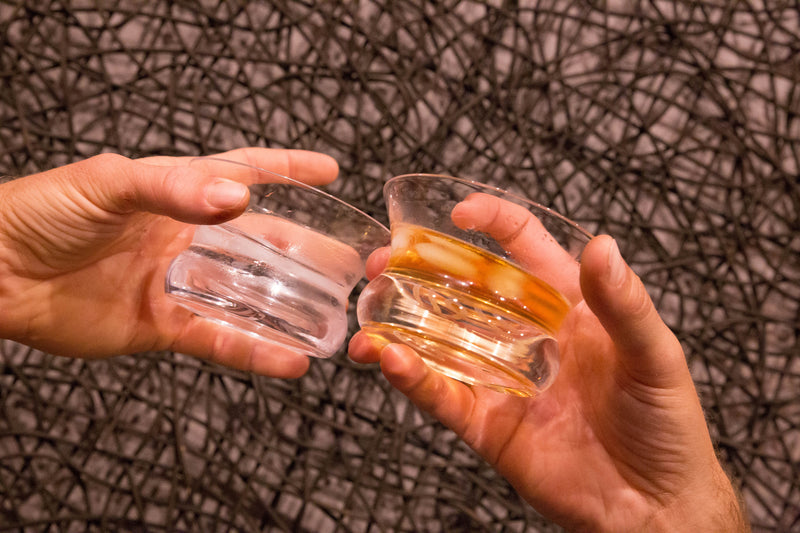 Two Oaxaca glasses being cheersed, filled with tequila and ice cubes causing condensation on the glass.