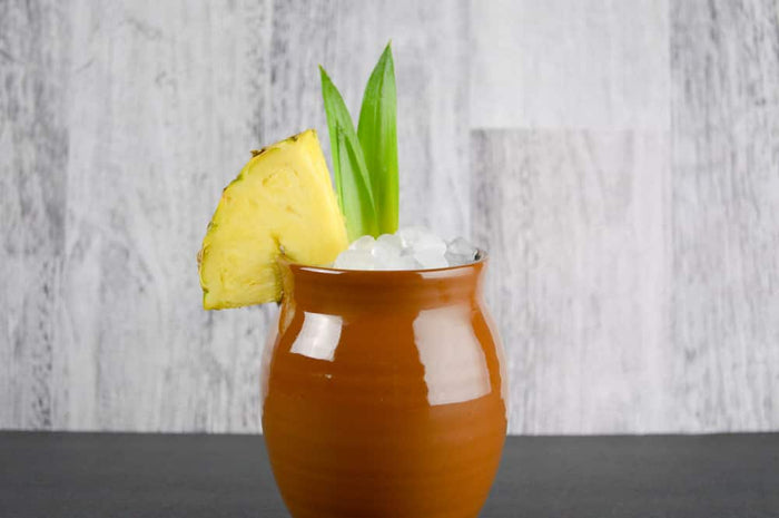 A Glossy finish Jalisco jug on a concrete countertop with a gray wood background. The jug is filled with small ice cubes and garnished with a pineapple slice and pineapple leaves.