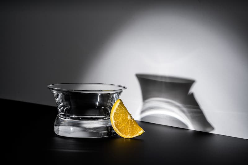 Oaxaca glass on black counter with a white background and an orange slice. The room is dark and the glass is lit from the left showing its shadow on the wall.