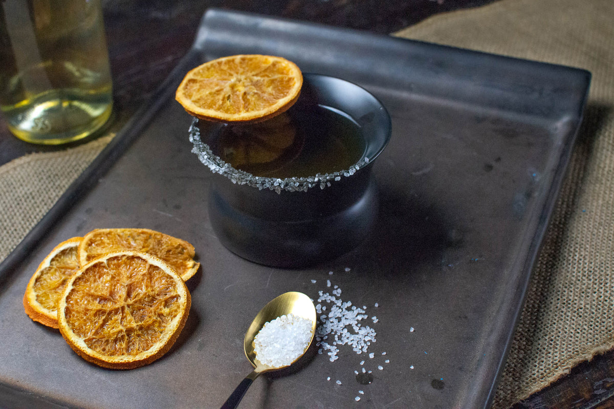 Volcanic ash (black) Oaxaca glass on a black tray filled with tequila, garnished with a salted rim and dried orange slices.
