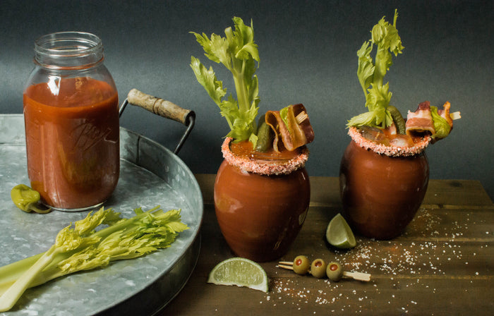 Two glossy finish Jalisco jugs sit on a wooden countertop next to a metal tray holding a canning jar full of Bloody Mary mix and other garnishes. The two jugs contain are rimmed with salt, and filled to bursting with celery stalks, skewered bacon, pickles, and jalapeños. Just a tiny bit of the Bloody Mary cocktail can be seen.