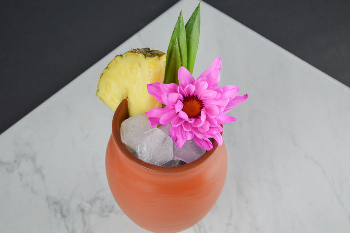 Single Jalisco jug on a marble countertop, filled with hexagonal ice cubes, garnished with a pineapple slice, a flower, and pineapple leaves.