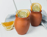 Two matte finish Jalisco jugs shown on a white countertop with a white backgroun, filled with a cocktail, red salted rim and citrus slices.