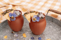 Two matte finish Jalisco jugs shown on a metal tray with cork handles, on a yellow gingham tablecloth. The jugs are filled with small ice cubes, and garnished with pineapple slices and flowers.